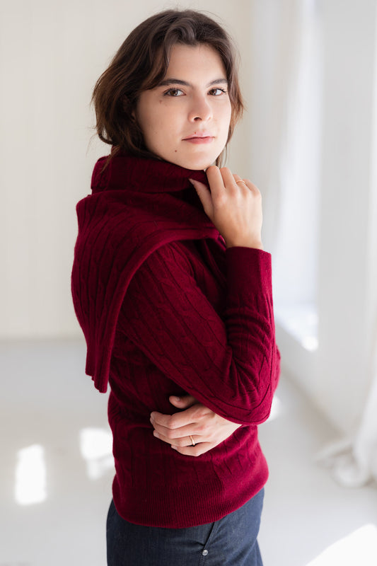 A woman wearing a deep burgundy cable-knit cashmere sweater and matching knit scarf, standing in a bright, minimalist room.