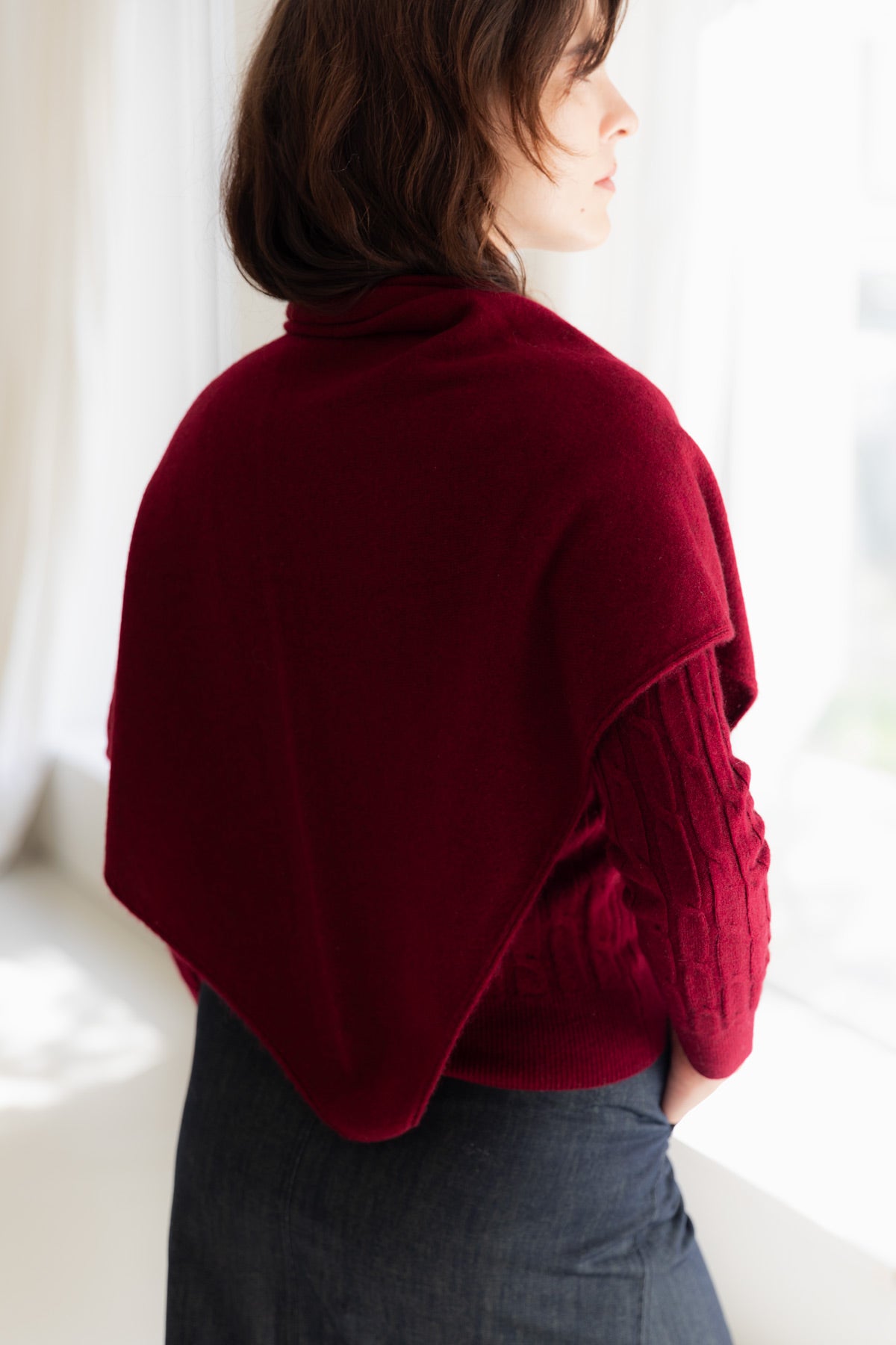 A woman shown from the back wearing a deep burgundy cashmere triangle scarf over a cable-knit cashmere sweater and dark denim skirt.