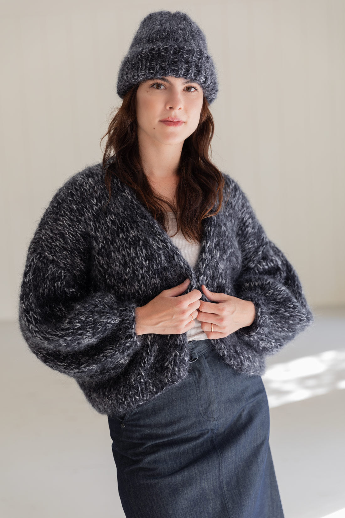 A woman wearing a chunky mohair beanie and a matching mohair cardigan, knitted in a dark mouilnee, standing against a plain light beige wall.
