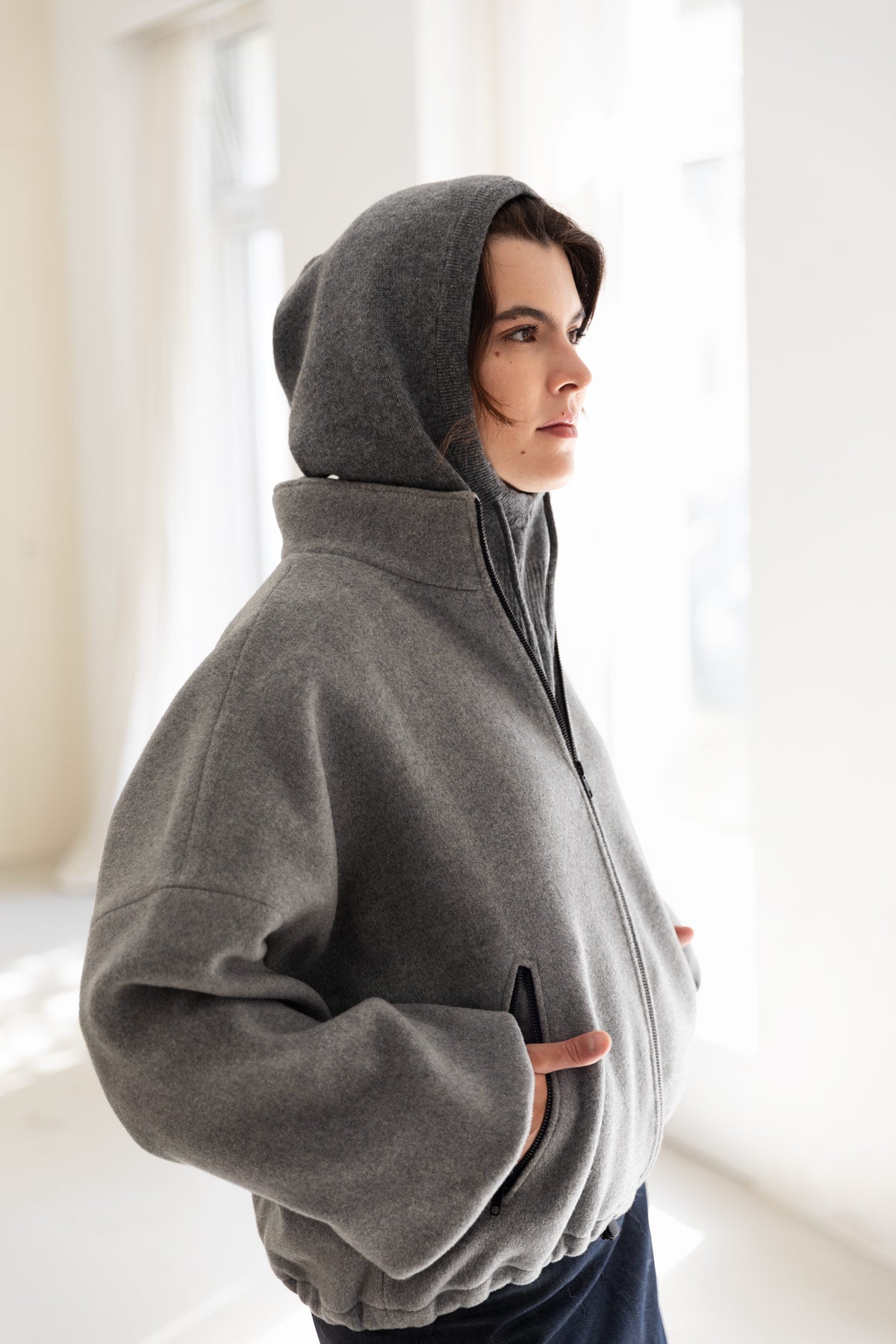 Woman wearing a grey cashmere balaclava, paired with a grey bomber jacket standing in a bright, minimalist room.