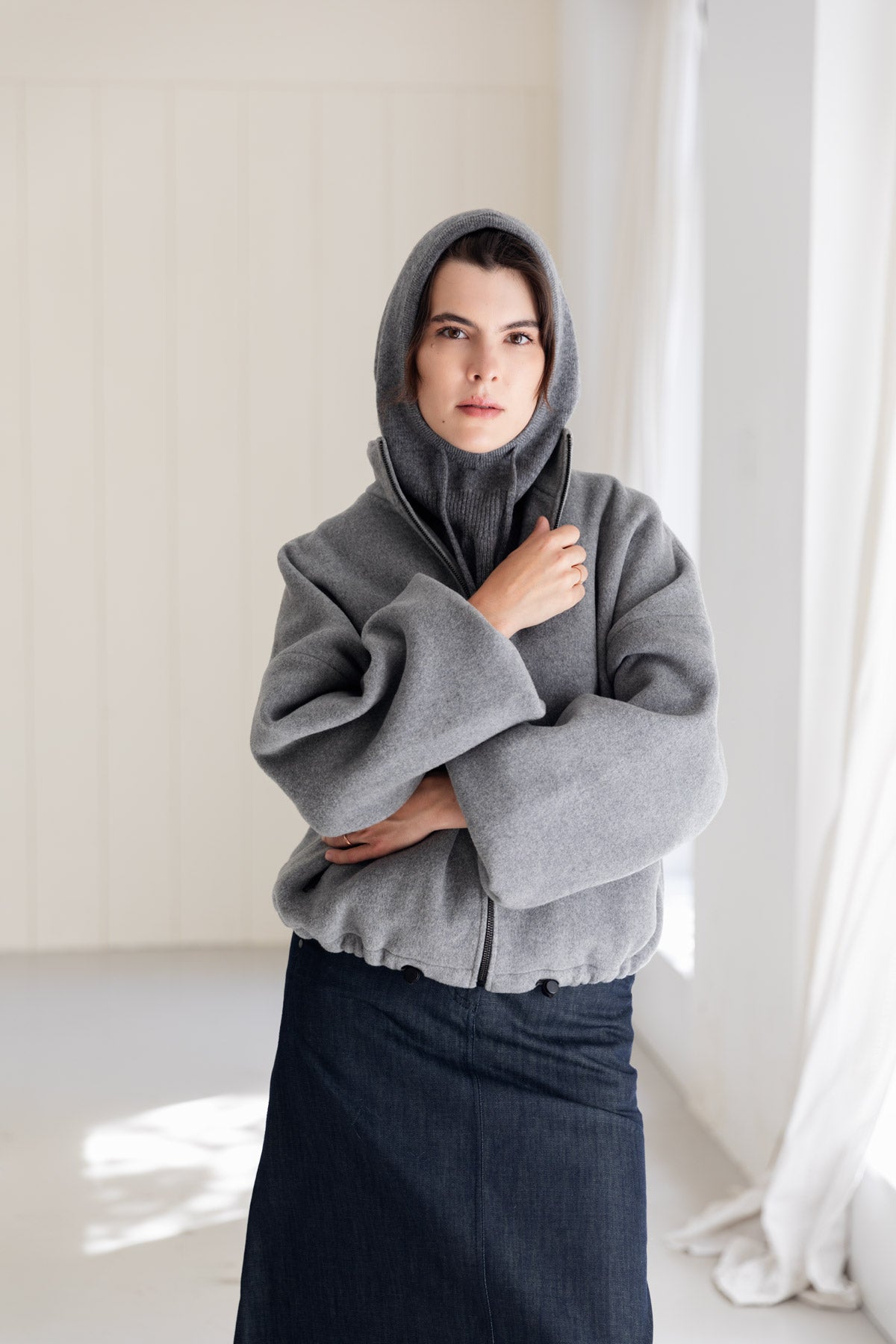 Woman wearing a grey cashmere balaclava, paired with a grey bomber jacket and denim skirt, standing in a bright, minimalist room.