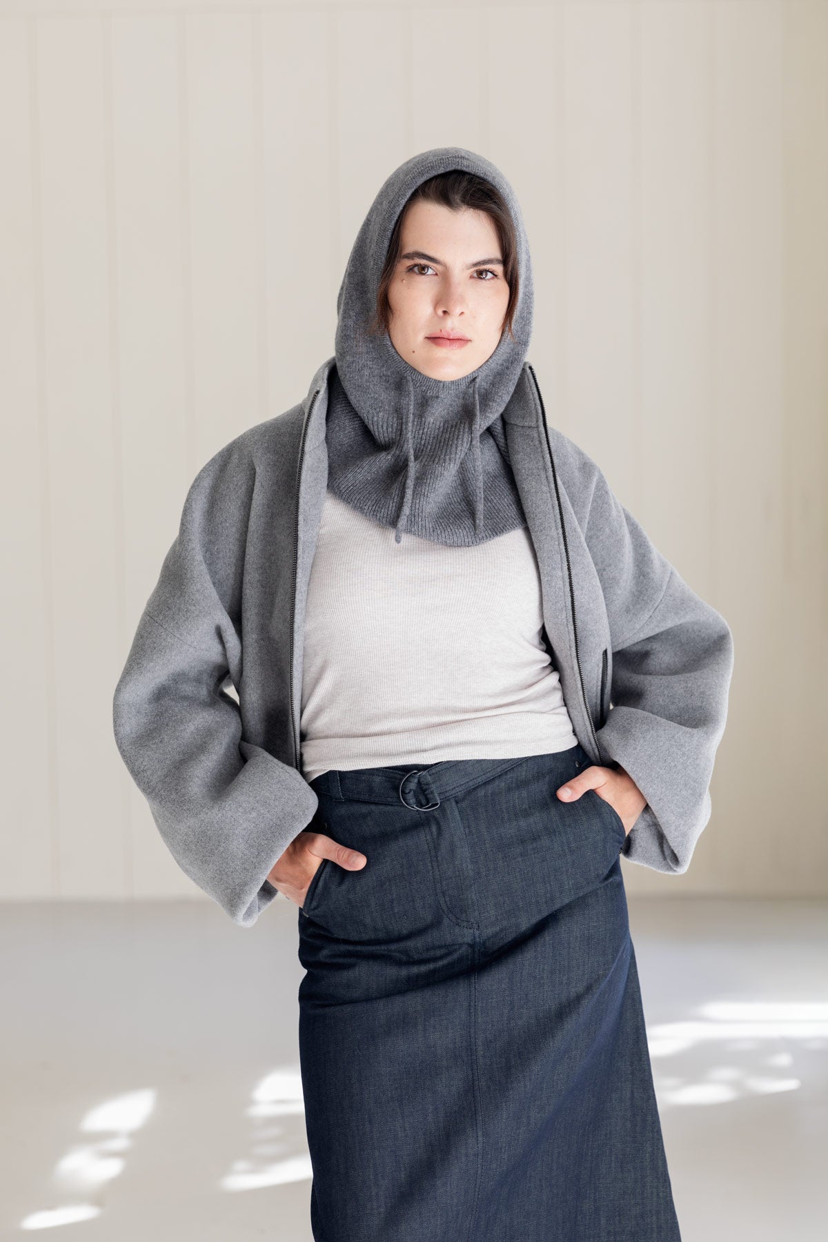 A woman wearing a grey cashmere balaclava, matching grey bomber jacket, and dark denim skirt.