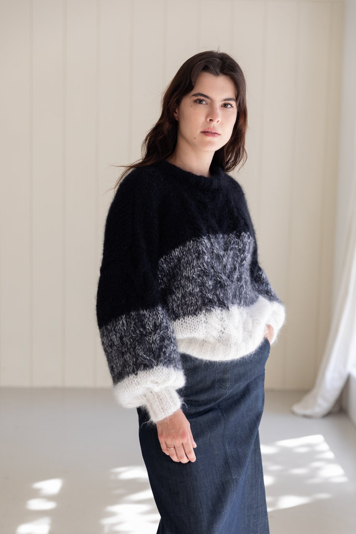 A woman wearing a fuzzy colour-block mohair sweater with black, grey, and white stripes, paired with a dark denim skirt.