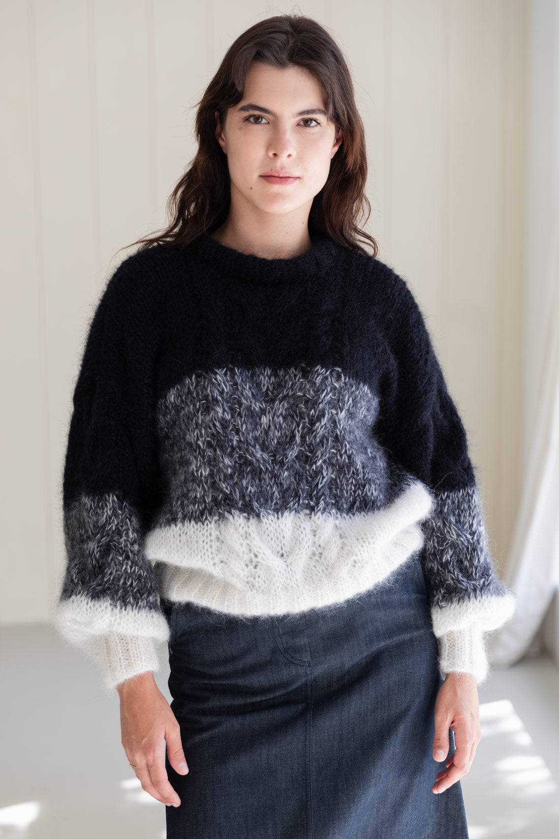 A woman wearing a fuzzy mohair sweater with a colour-blocked pattern in black, grey, and white, paired with a dark denim skirt.