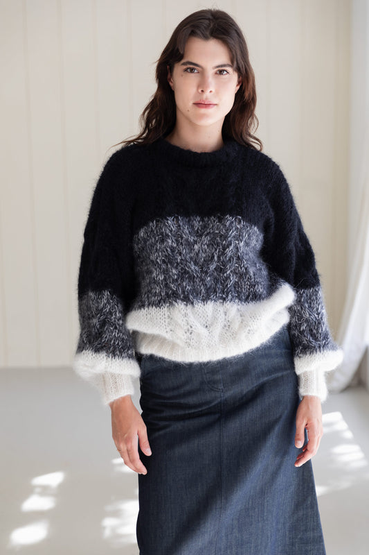 A woman wearing a mohair sweater with horizontal colour-blocked stripes in black, grey, and white, paired with a dark denim skirt, standing in a brightly lit indoor setting.