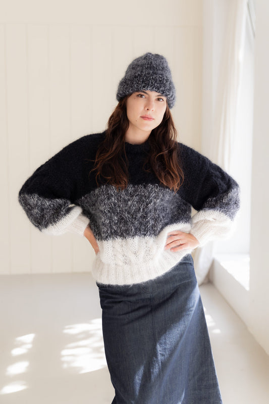 A woman wearing a chunky mohair sweater with black, grey, and white stripes, a matching mohair beanie, and a dark denim skirt, standing in a bright, sunlit room.