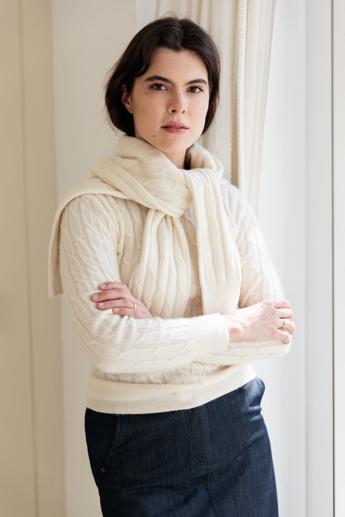 A woman wearing a cream cable-knit cashmere sweater, paired with dark denim skirt, standing in front of a soft beige curtain.