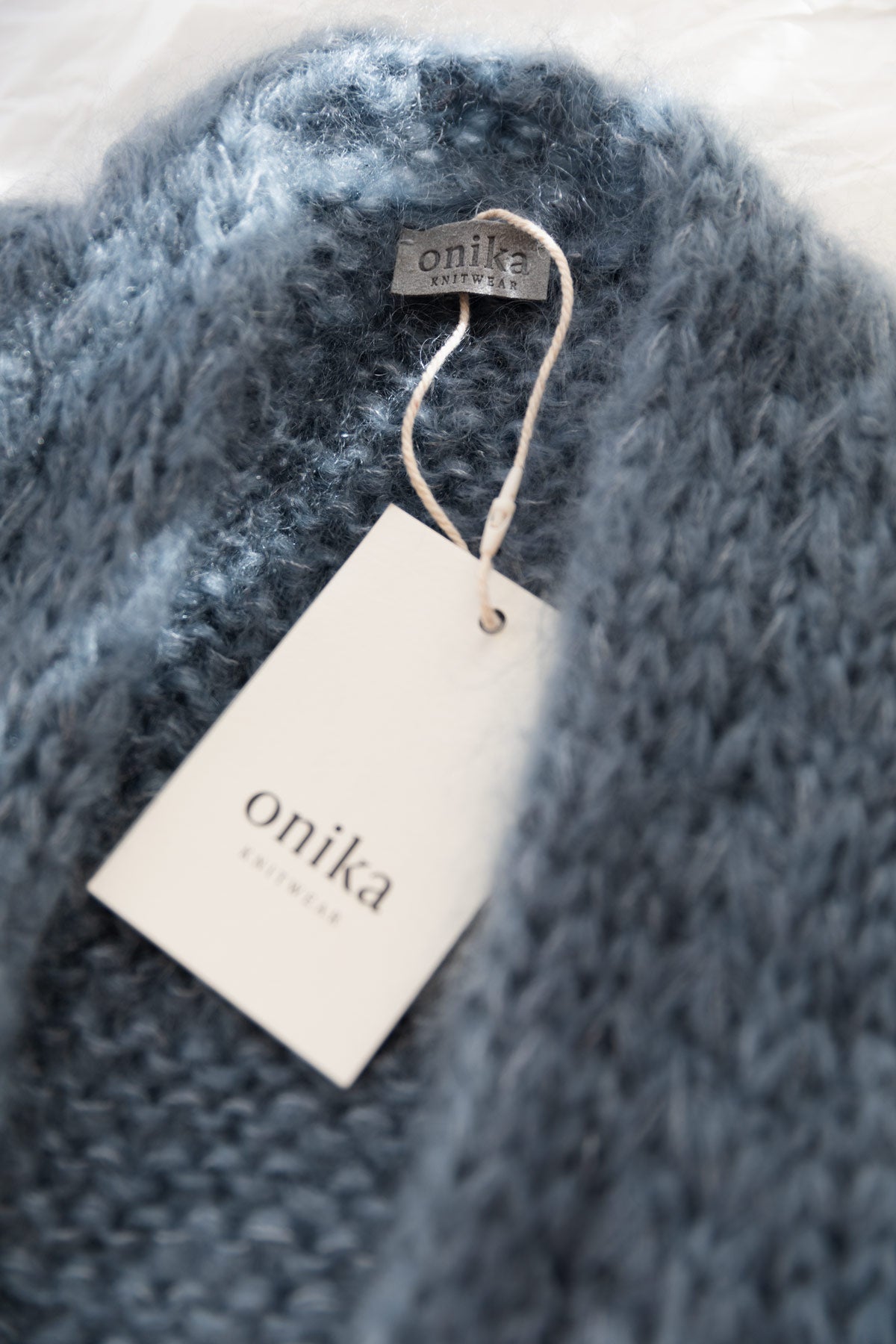 A close-up of a soft, fuzzy mohair cardigan in a muted blue-grey colour, featuring a white paper tag and a small brand label that reads 'onika knitwear'.