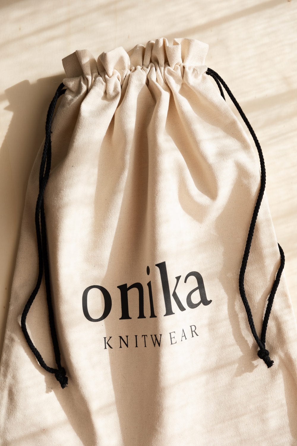 A cream cotton dust bag with black cords and the brand name 'onika KNITWEAR' printed in black on the front, photographed on a light wood surface with soft shadows.