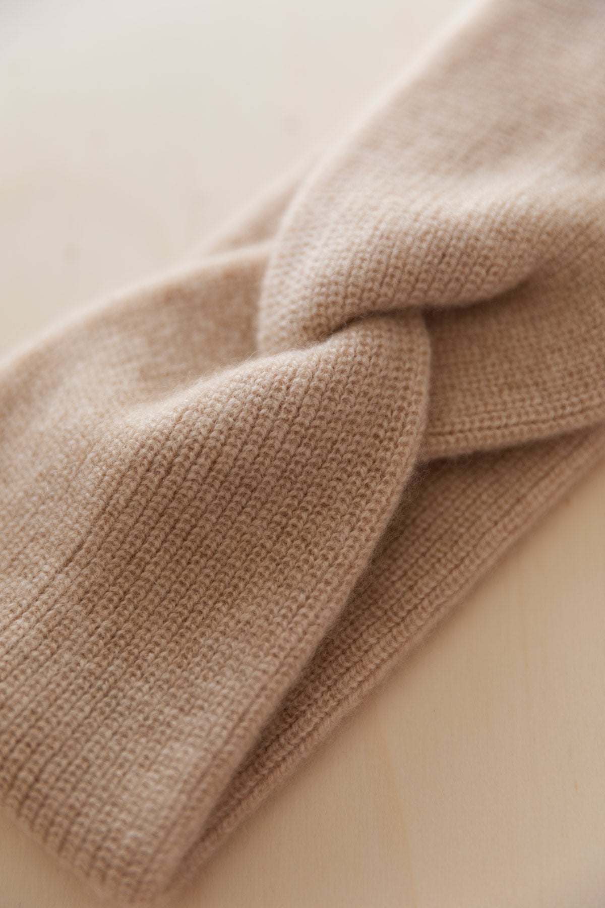 A close-up of a beige cashmere headband with a twisted knot detail, shown on a light beige background.