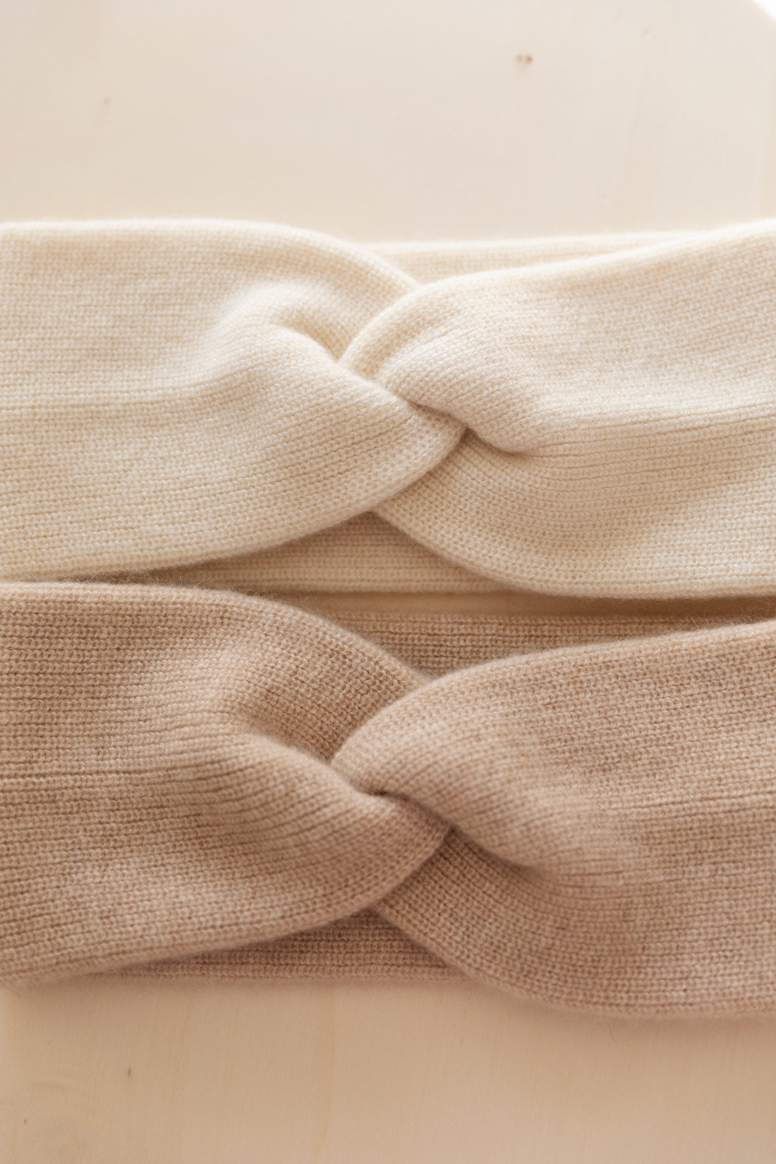 A close-up of two cashmere headbands, with a twisted knot detail, in white and beige, shown on a light beige background.