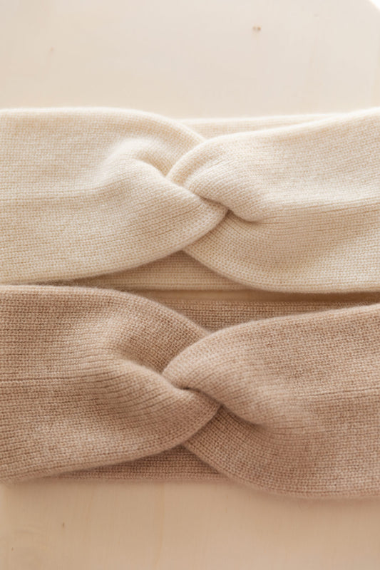 A close-up of two cashmere headbands, with a twisted knot detail, in white and beige, shown on a light beige background.