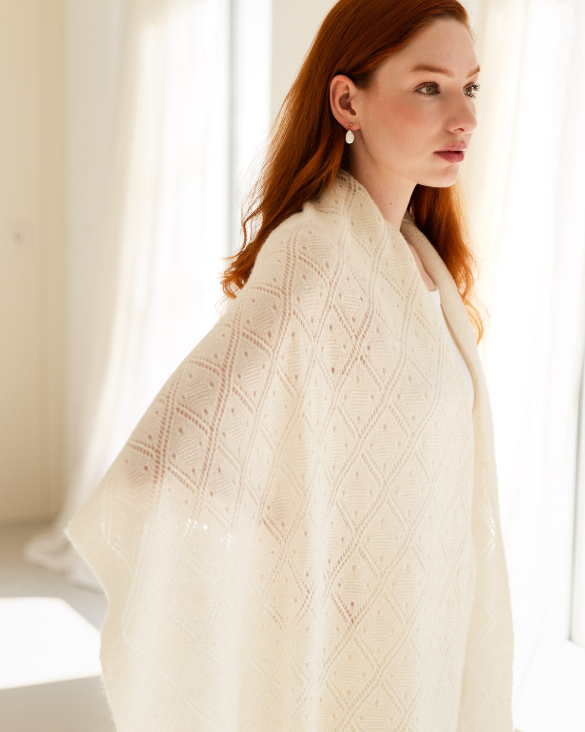 A woman with red hair wearing a cream lace cashmere shawl, standing in a brightly lit room with soft window light.